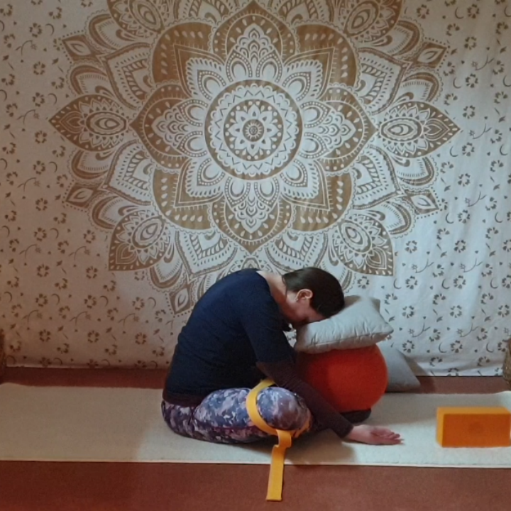 Marion übt Yin Yoga und übt die Position vorgebeugter Schmetterling. Die Fußsohlen sind aneinandergelegt, ein Yogagurt ist um die Knie gelegt und stützt den Bereich. Die Stirn ist abgesenkt und ruht auf zwei Kissen.