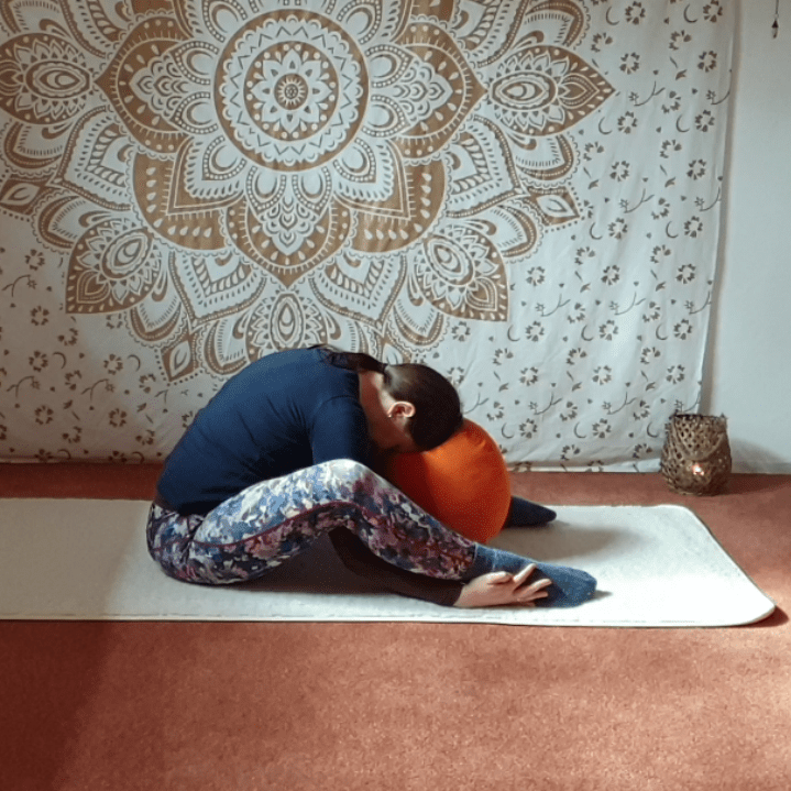 Marion übt Yin Yoga und die Position Kutscher. Sie sitzt, die Füße sind auf der matte aufgesetllt. Der Oberkörper ist vorgebeut und die Stirn ruht auf einem Yogakissen. Die Hände umfassen die Knöchel an der Außenseite.